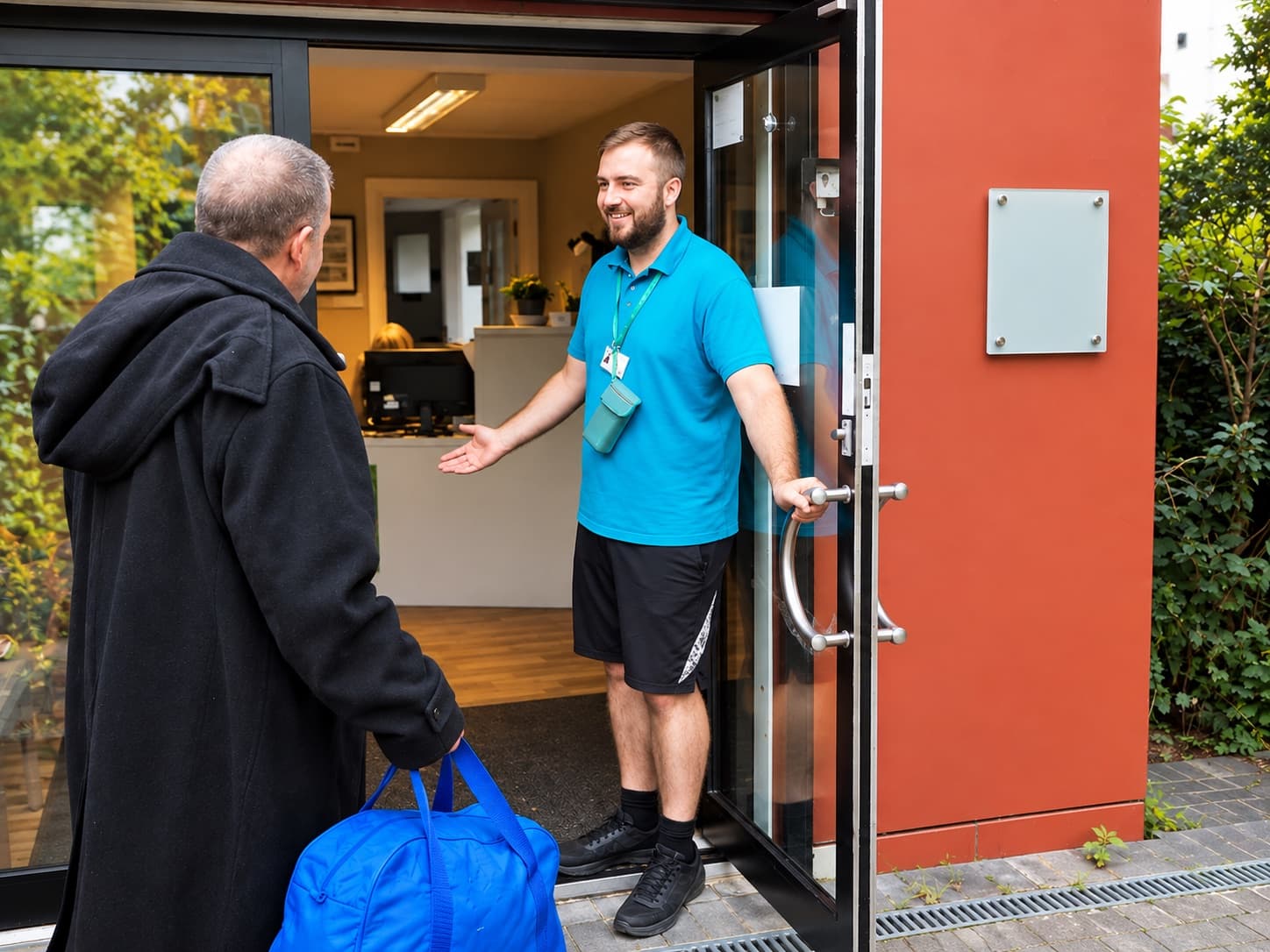 A smiling shelter worker holds the door open and welcomes a man carrying a bag